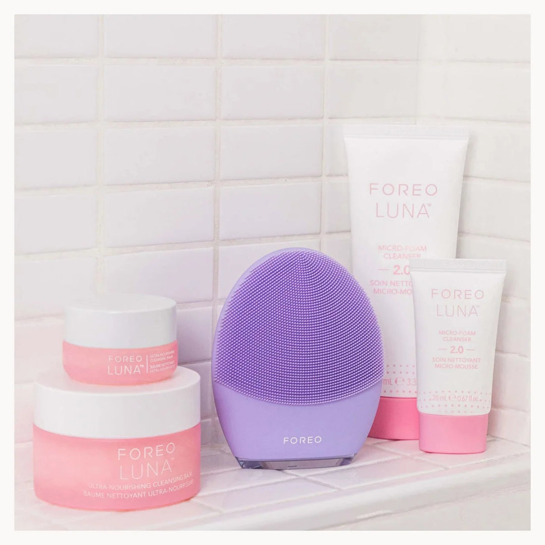 A Foreo Luna facial cleansing device in purple stands on a tiled bathroom shelf alongside several Foreo Luna skincare products in pink and white packaging.