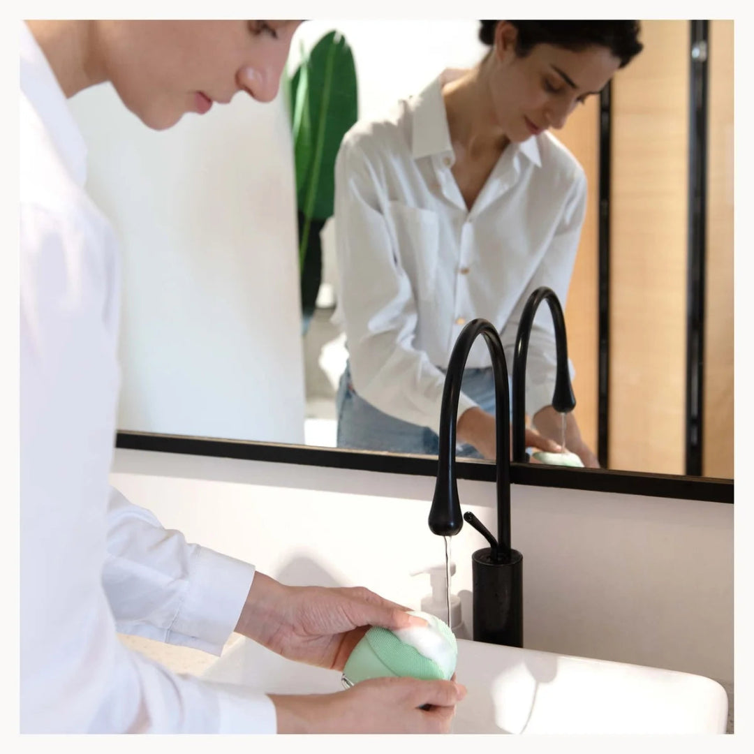 A person in a white shirt washes a mint green and white oval soap bar in a white sink, with a black faucet and their reflection visible in a mirror.