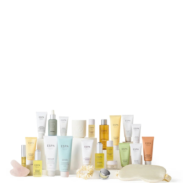A collection of ESPA skincare and wellness products arranged on a white surface against a white background, including tubes, bottles, a mask, a sleep mask, and small accessories in soft pastel and yellow tones.