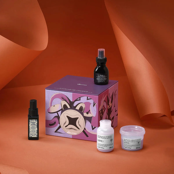 A curated beauty set featuring a purple illustrated gift box, a black spray bottle, a small black dropper bottle, a white bottle labeled SAFE!, and a clear jar with white lid, arranged on a warm terracotta background with draped paper folds.