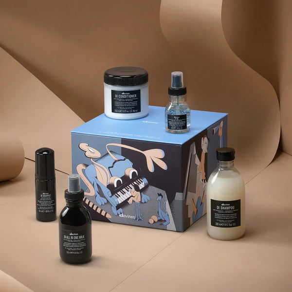 A boxed haircare set from divines displayed on a beige sculptural backdrop. The set includes a blue illustrated gift box with cartoon-style artwork, a white conditioner jar, a clear bottle with blue liquid and spray top, a black bottle, and a frosted bottle with creamy shampoo.