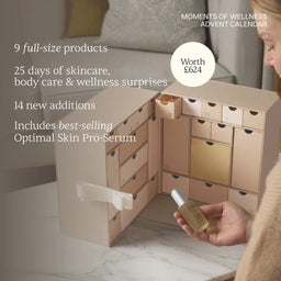 A beige advent calendar box with multiple numbered drawers is shown open on a marble surface in a softly lit indoor setting. A person's hand holds a small skincare bottle in front of the calendar, and promotional text on the left reads about wellness products and value.