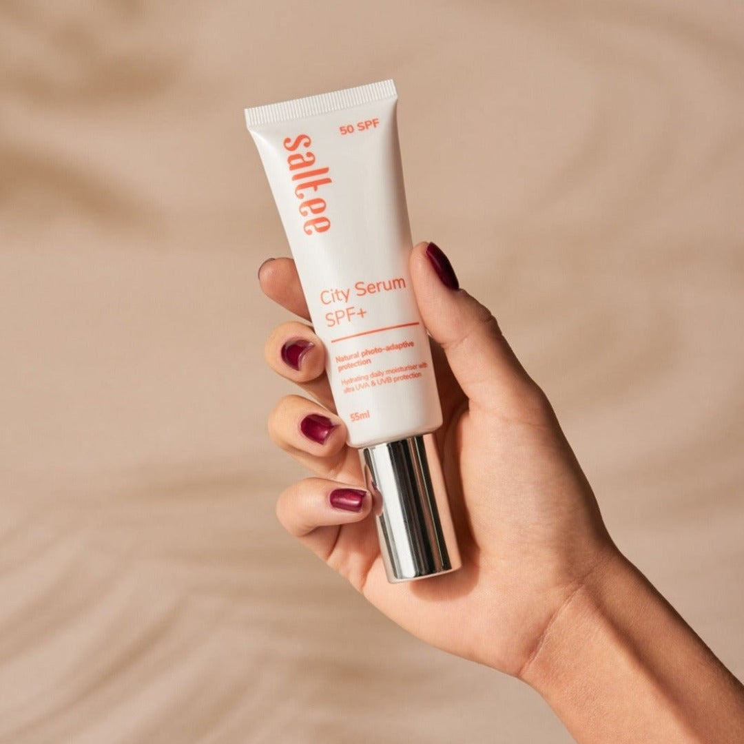 a hand holding a tube of Saltee City Serum SPF+ on a sandy beach